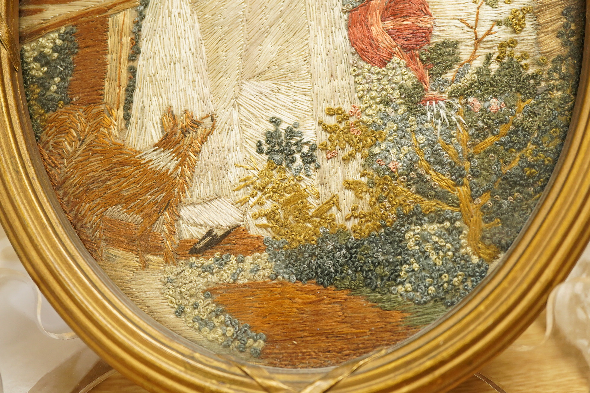 A small oval gilt framed late 19th century silk polychrome embroidery of a lady watering her garden with her dog at her side, 19cm high. Condition - good.